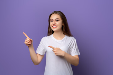 Portrait of young woman pointing fingers aside and looking with positive face expression, standing over purple background