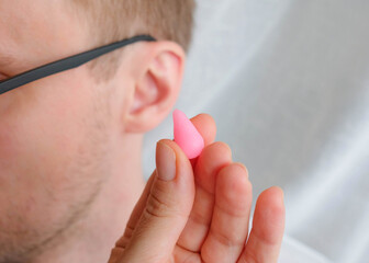 Holds a pink wax earplug in his hand. Human ear in the background