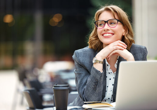 Lovely  businesswoman in glasses sitting with laptop and cup of takeaway coffee outside