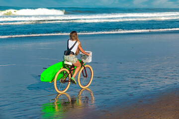 Young woman with surfboard and bicycle on the beach.