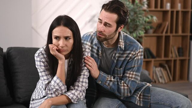 Candid caucasian young man consoling his upset wife or girlfriend, while sitting on couch at home in living room together, they experiencing difficulties in relationships or or personal problems