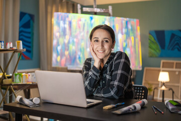 Portrait of smiling brunette wearing a checkered shirt sitting at a desk in front of a laptop in...