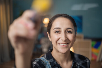 Portrait of a smiling, happy lady stretching out her hand in which she holds a brush dirty with yellow oil paint, watercolor, in the background living room in the evening, canvases, easels