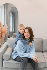 A young mother and a little son spend time at home. They sit in the living room on the sofa and hug.