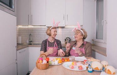 two happy elderly women with rabbit ears on their heads are painting Easter eggs at home in the kitchen. Preparation for the Easter feast