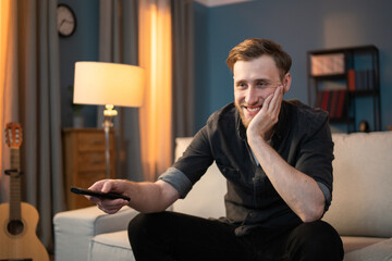 A smiling boy sits on couch in evening in living room, holding TV remote in hand, switching channels, looking for a movie to watch, propping hand under chin, face, showing teeth