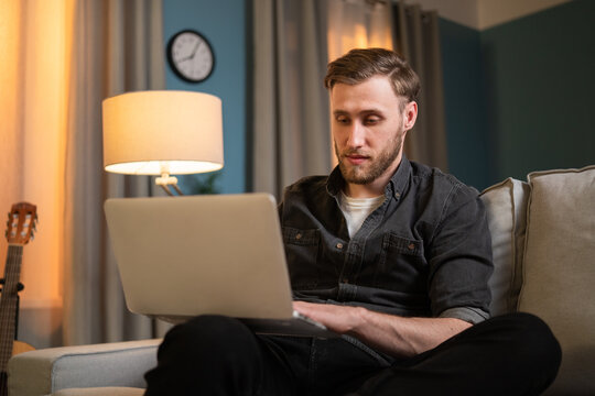 Entrepreneurial guy works late in home office sitting on couch, man holds computer, laptop on lap, takes out insurance for his business, pays bills, answers emails, does online shopping
