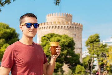A happy smiling young man drinks traditional Frappe coffee against the background of the famous...