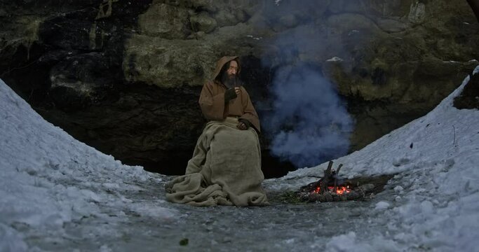 Druid smokes tobacco pipe warming near bonfire against cave