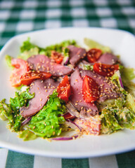 Healthy green salad with meat, tomato and lettuce served in a square white plate over green plaid tablecloth