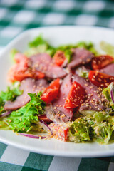 Healthy green salad with meat, tomato and lettuce served in a square white plate over green plaid tablecloth