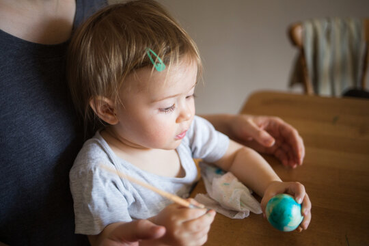 baby child paints Easter eggs with mother at home in kitchen. kids craft.