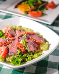 Healthy green salad with meat, tomato and lettuce served in a square white plate over green plaid tablecloth