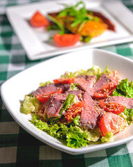 Healthy green salad with meat, tomato and lettuce served in a square white plate over green plaid tablecloth