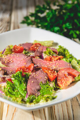 Healthy green salad with meat, tomato and lettuce served in a square white plate over grey or gray background.