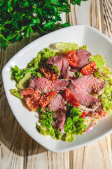 Healthy green salad with meat, tomato and lettuce served in a square white plate over grey or gray background.