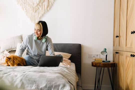 Asian girl petting her dog while using laptop on bed - Powered by Adobe