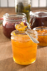 A spoon of marmelade on top of a jam jar, with strawberry and other jams in the background.