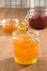 A spoon of marmelade on top of a jam jar, with strawberry and other jams in the background.