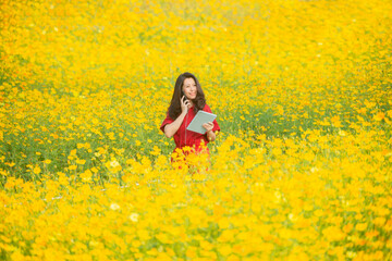 Fototapeta premium Asian farmer woman talking on phone and using tablet for checking report, collect data to study of agriculture in flower farm. Blurred background of yellow cosmos flower field. Copy space