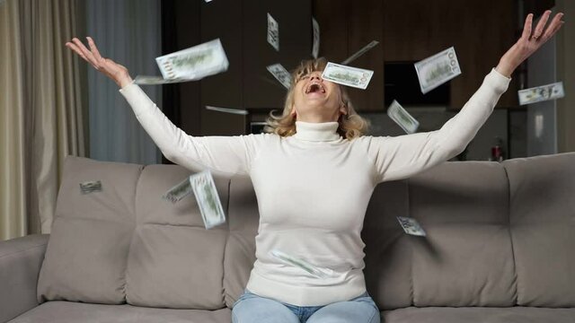 Blonde aged dame with bangs wearing turtleneck tosses cash banknotes of pension into air of living room smiling and laughing happily closeup slow motion.