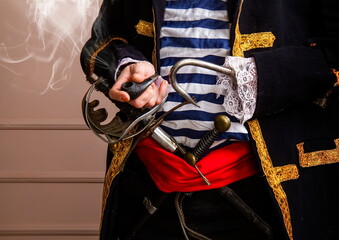 A brave armed pirate captain in a frock coat and a vest with a sword and a dagger and a hook smokes a pipe close-up