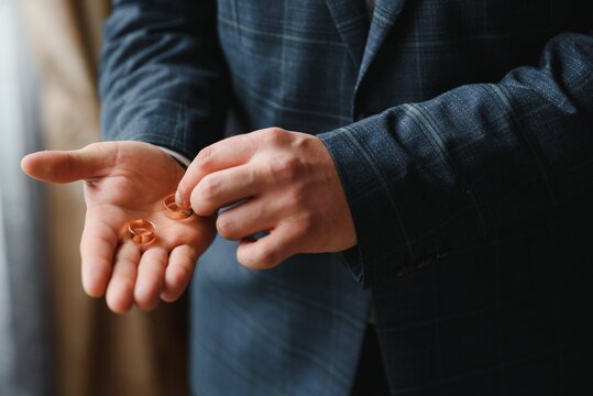 The groom on his wedding day holds two wedding rings in his hand in a hotel room. The man is wearing a white shirt and blue trousers with a vest. Male hand and wedding rings close up