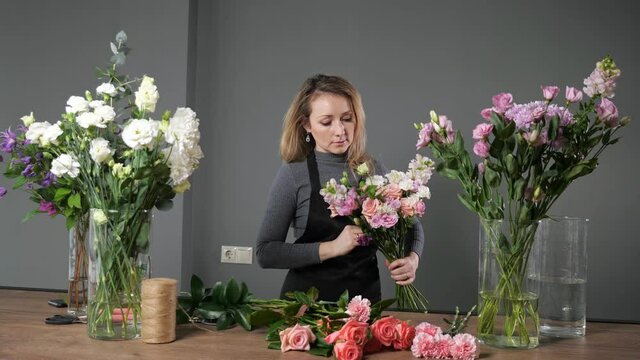 Florist blonde woman in black apron composes elegant spring bouquet with colorful flowers and green twigs on wooden table in flowershop.