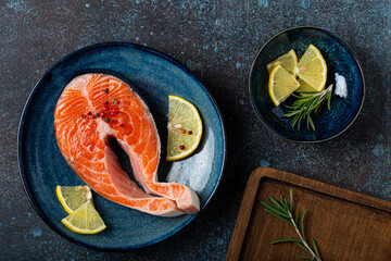 Uncooked raw fresh fish salmon steak top view on plate rustic dark concrete stone background with rosemary, lemon wedges and spices, delicacy healthy fish cooking and nutrition concept flat lay