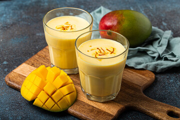 Healthy Indian Ayurveda drink mango lassi in two glasses on rustic concrete table with fresh ripe cut mango, yellow blended beverage made of mango fruit, yoghurt or milk curd and spices 