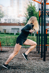 Woman in workout. Young athletic female in sportswear trains with fitness straps on sports ground.