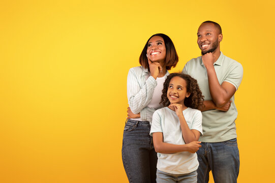 Special offer. Young black family of three looking aside at free space and holding hands on chin, yellow background