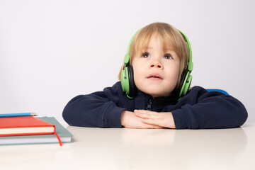 Close up portrait of cute little boy wearing headphones sitting at the table and looking up isolated over white background.