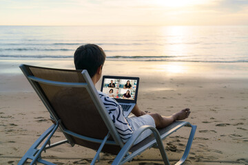 Asian business man having remote video conference call with his business team at the beach during vacation in holiday..