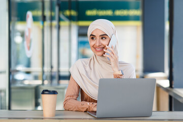 Smiling friendly cute millennial arab muslim woman in hijab speaks on phone and types on laptop in...