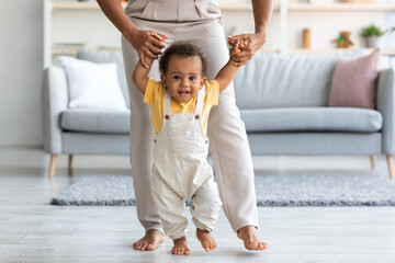 Adorable black infant boy making first steps at home with mother's help