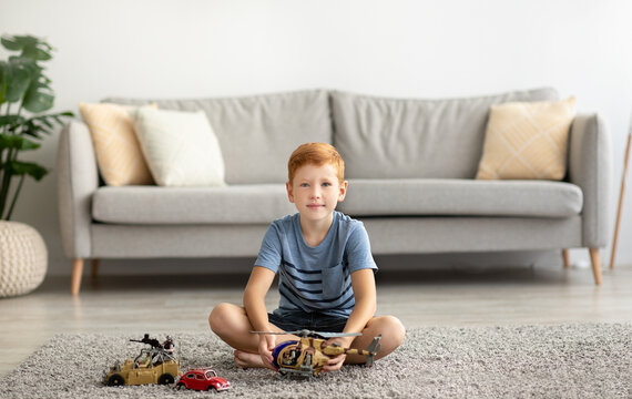 Cheerful boy sitting on carpet, playing with helicopter and cars