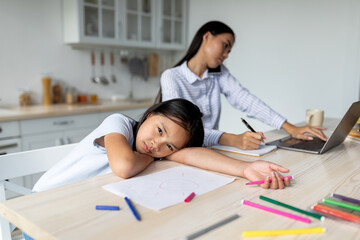 Young korean mother working from home, ignoring her offended little daughter, girl feeling lonely