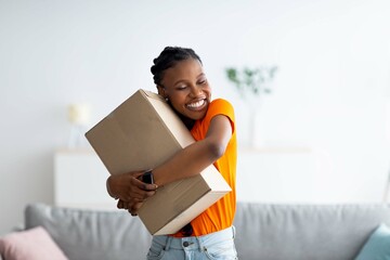 Cheerful Afro woman hugging carton parcel, receiving long awaited delivery, getting online order indoors - obrazy, fototapety, plakaty