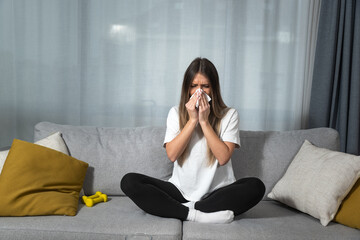 Young woman suffering from a common cold and flu or allergy sitting at home and wipe her nose with tissues while she have strong headache pain. Female with strong allergic reaction healthcare concept - obrazy, fototapety, plakaty