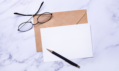 On a light marble background, a craft envelope, glasses, a pen and a white card with a place to...