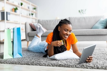 Black lady lying on soft carpet with tablet pc and credit card, surrounded by gift bags, shopping...