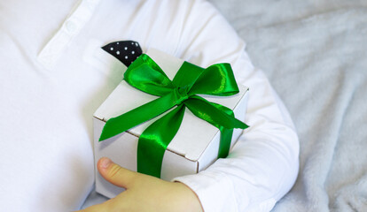 handsome brunette hair boy is hugging a white gift box with green color ribbon in his hands. cute kid fall asleep, in white shirt with black bow, ready for celebration. woman, mother's day.