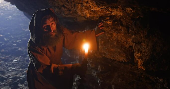 Old druid walks carefully in dark ancient cave with candle