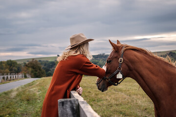 Cowgirl stroking her horse at ranch. Woman with cowboy hat at pasture in animal farm. Friendship between people and horse - obrazy, fototapety, plakaty