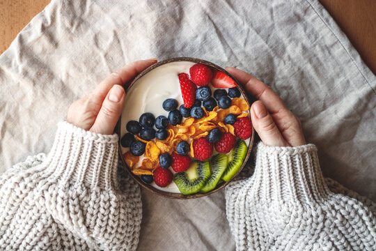 Healthy breakfast in coconut bowl on table with linen tablecloth. Yogurt with corn flakes and blueberry, strawberry, kiwi and raspberry. Womans hand wearing sweater. Vegetarian food 