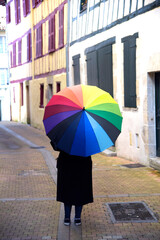 Una persona con ropa negra y un paraguas con los colores del arcoiris de espaldas paseando por una calle de un pueblo frances.