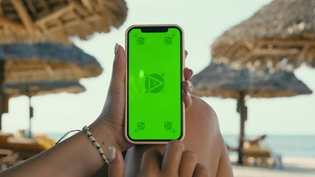 Mockup of a Young Woman Holding Phone with Green Screen Laying on a Sun Lounger on the Beach. Mobile Smartphone App Commercial Concept. X2 Slow Motion