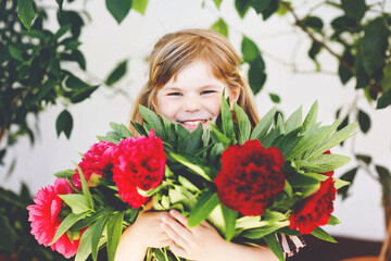 Cute adorable little toddler girl with huge bouquet of blossoming red and pink peony flowers. Portrait of smiling preschool child in domestic garden on warm spring or summer day. Summertime.