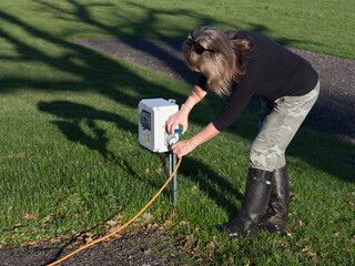  A female motorhomer plugs the orange electric hook up lead into a campsite supply point using the blue connector.She wears rubber boots and camouflage trousers on sunny day with sunglasses on head - obrazy, fototapety, plakaty